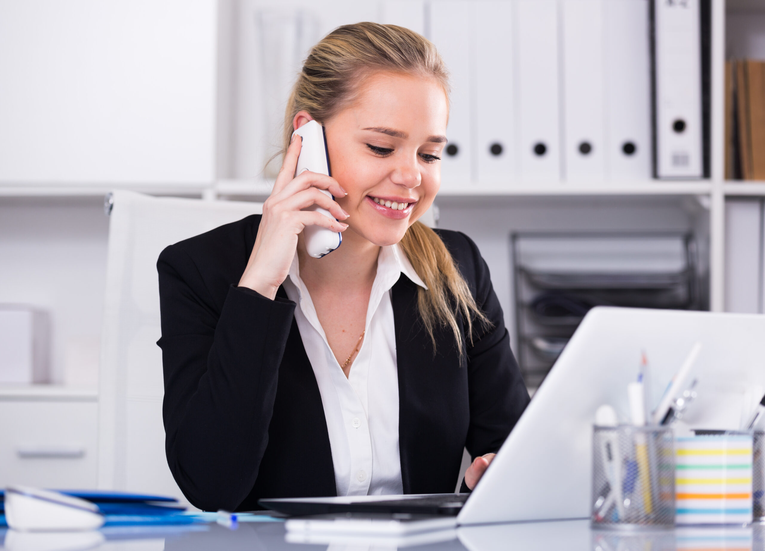 Woman talking on phone in office Smiling woman talking on mobile phone and using laptop in office